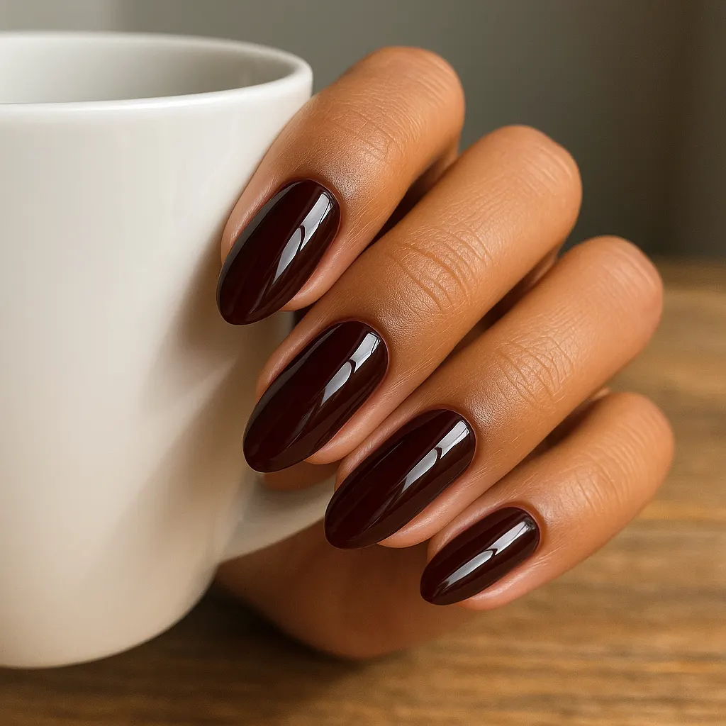 Medium almond nails in a glossy cherry mocha, a red-brown so deep it reads nearly black, on warm tan skin holding a white mug.