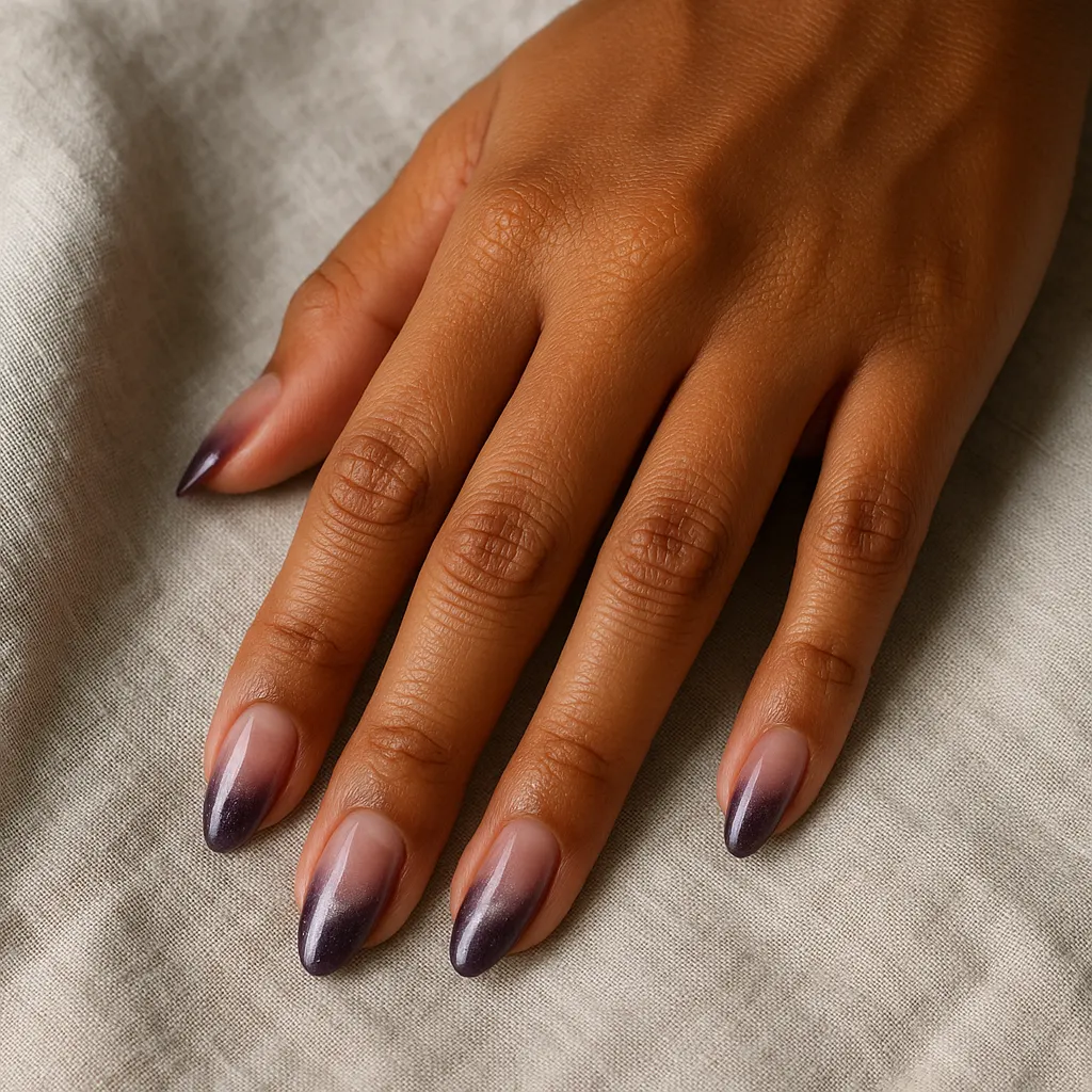 Medium oval nails fading from sheer nude pink to deep plum tips with a faint shimmer, on morena skin resting on grey linen.