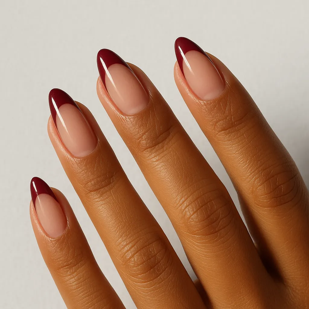 Medium almond nails, a nude base with a deep wine red tip, on morena skin against a light grey background.
